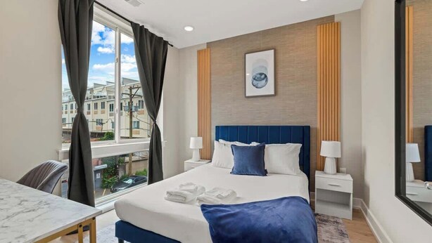 A hotel room with a large bed, a desk, and a view of the city through the window. - Luxury 1BD | 2 Beds | Northern Liberties (Philadelphia)