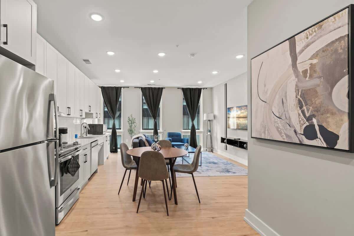 A modern kitchen with stainless steel appliances, a dining area with a round table and chairs, and a large abstract painting on the wall.