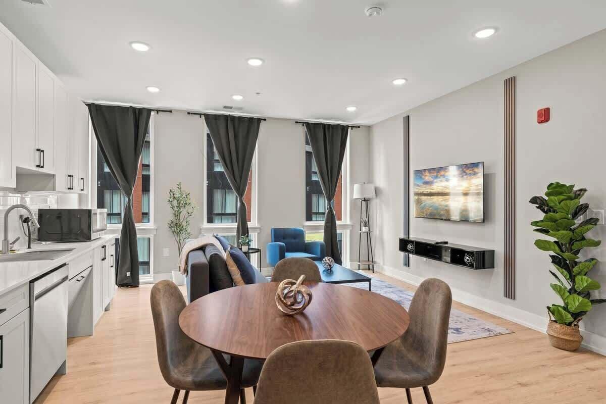 A modern kitchen with white cabinets, a microwave, and a sink. There is a dining area with a round table and four chairs. A blue sofa and a lamp are visible in the background.