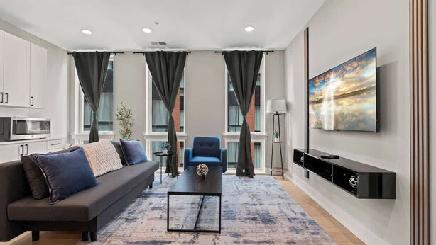 A modern living room with a sofa, a coffee table, a TV mounted on the wall, and a view of the outside through large windows.