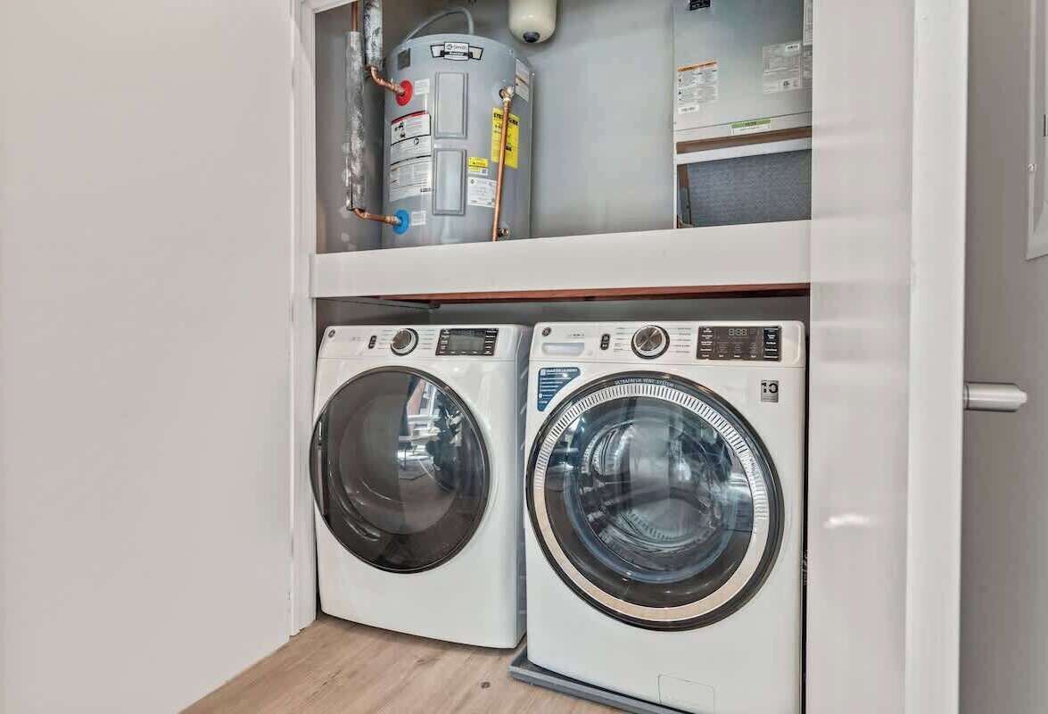 A laundry room with two front-loading washing machines and a water heater on the shelf above.