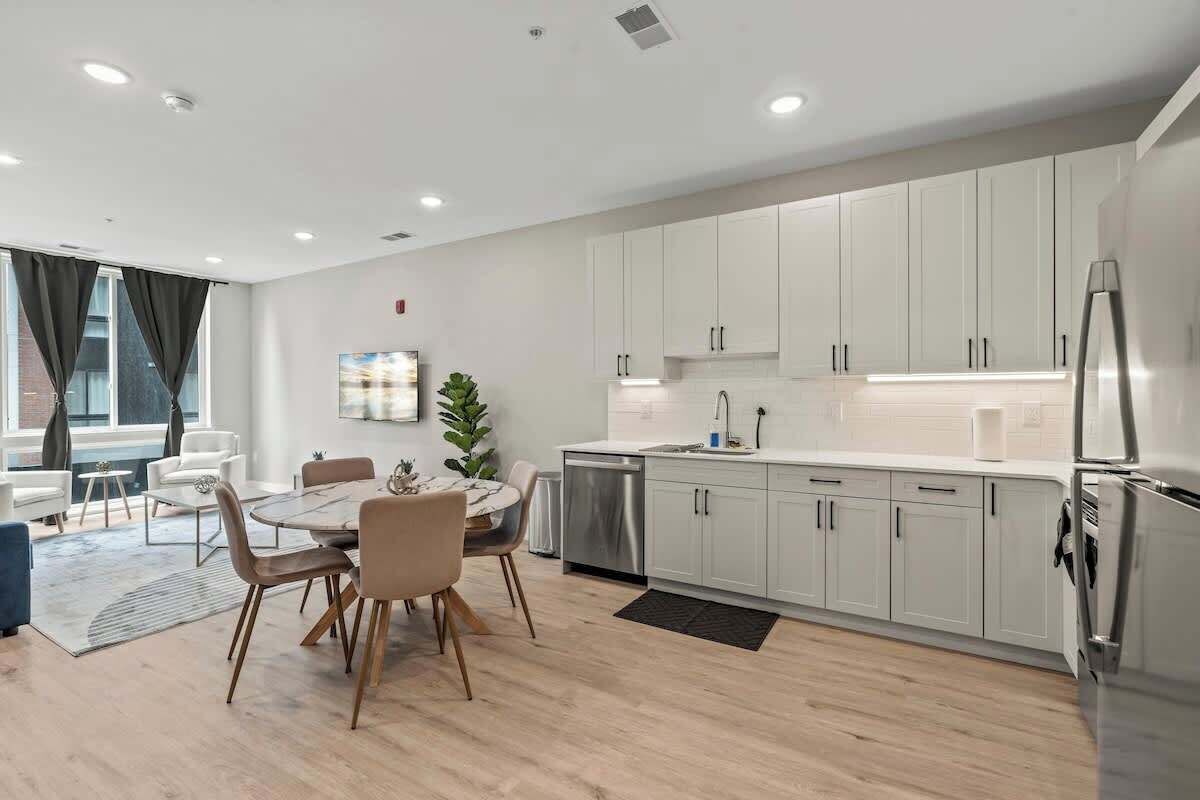 A modern kitchen with white cabinets, a stainless steel refrigerator, and a dining area with a round table and chairs.