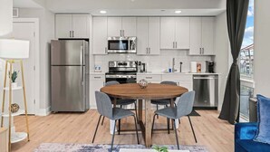 A modern kitchen with stainless steel appliances, a dining area with a wooden table and grey chairs, and a view of a cityscape through large windows.