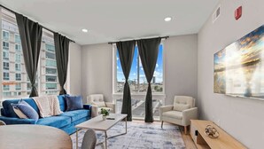 A modern hotel room with a blue sofa, a white armchair, a wooden coffee table, and a large window with curtains.