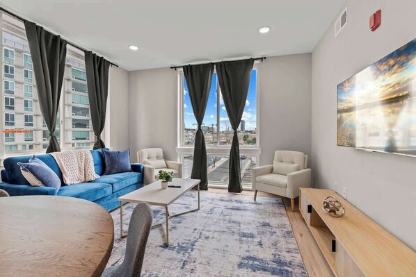 A modern hotel room with a blue sofa, a white armchair, a wooden coffee table, and a large window with curtains.
