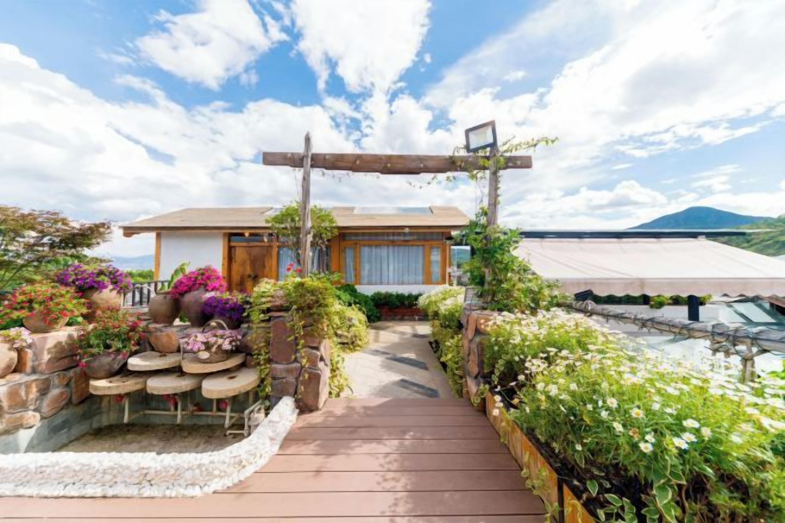 Une maison traditionnelle de style japonais, dotée d’une terrasse en bois menant à l’entrée, et entourée d’une végétation luxuriante et de fleurs.