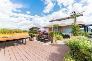 A wooden deck with a table and chairs, surrounded by lush greenery and a small water feature.
