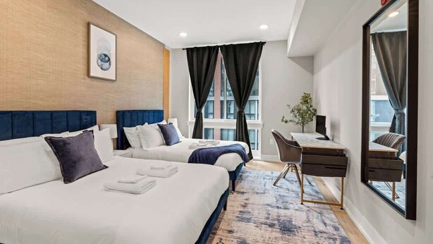Ein Hotelzimmer mit zwei Betten, einem Schreibtisch, einem Sessel und einem großen Spiegel, sowie einem Fenster mit Vorhängen.