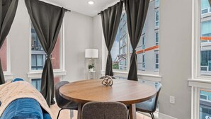A modern living room with a round wooden table, two chairs, a blue sofa, and large windows with dark curtains. - Luxury 1BD | 2 Beds | Northern Liberties (Philadelphia)