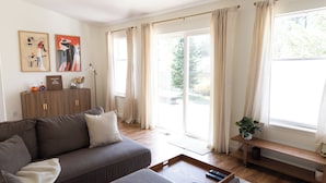 Ein modernes Wohnzimmer mit grauem Sofa, einem Holztisch und einer Bank am Fenster. Zwei gerahmte Kunstwerke hängen an der Wand und auf der Bank steht eine kleine Pflanze.