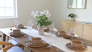 Ein Esstisch ist gedeckt mit Tellern, Schüsseln und Besteck. Eine Vase mit Blumen steht darauf, im Hintergrund ist ein Holzschrank zu sehen.