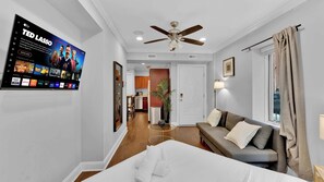 Quarto de hotel moderno com cama, sofá, ventoinha no teto, televisão exibindo um canal esportivo e um corredor que leva a uma cozinha.