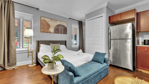 Ein modernes Hotelzimmer mit Bett, einer Couch, einem kleinen Tisch mit Pflanze, einer Küchenzeile mit Kühlschrank und einem großen Fenster mit Vorhängen.