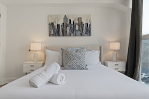 Une chambre d’hôtel avec un lit, deux tables de chevet avec des lampes, un tableau représentant un paysage urbain accroché au mur, et une fenêtre donnant sur une vue panoramique.