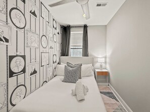 Un dormitorio con cama, mesita de noche, lámpara, ventana con cortinas y un cuadro con retratos en la pared.
