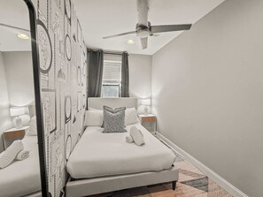 Une chambre à coucher avec un lit, deux tables de chevet, un ventilateur de plafond et une fenêtre avec des rideaux.