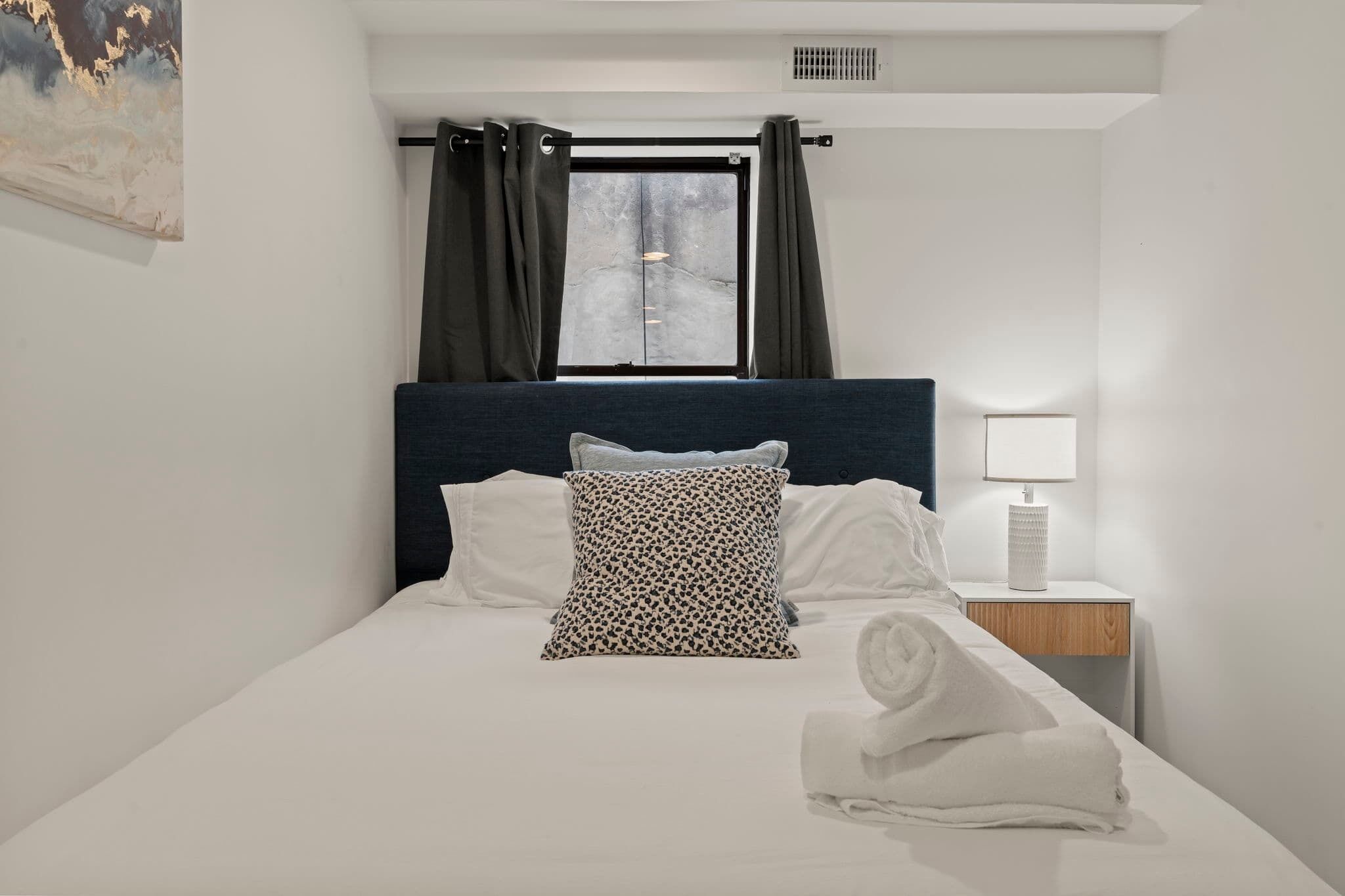 Una cama bien hecha con sábanas blancas y un cojín estampado de leopardo, una lámpara de noche y una ventana con cortinas.