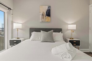 A neatly made bed with white linens, two bedside tables with lamps, a gray headboard, and a painting on the wall.