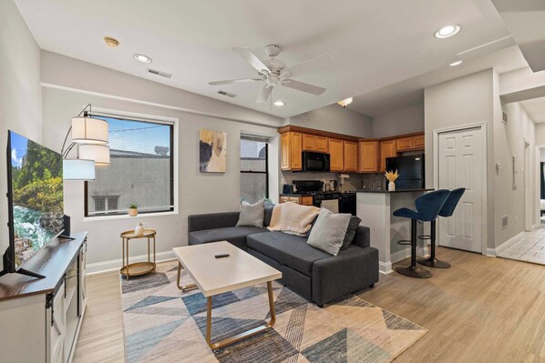 A modern living room with a sofa, coffee table, and TV. It includes a kitchen area with wooden cabinets and a ceiling fan.