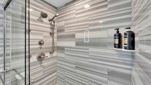 Une salle de bain moderne avec une douche à l’italienne en verre, des carreaux de marbre et deux bouteilles de shampoing posées sur une étagère.