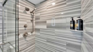 Une salle de bain moderne avec une douche à l’italienne en verre, des carreaux de marbre et deux bouteilles de shampoing posées sur une étagère.