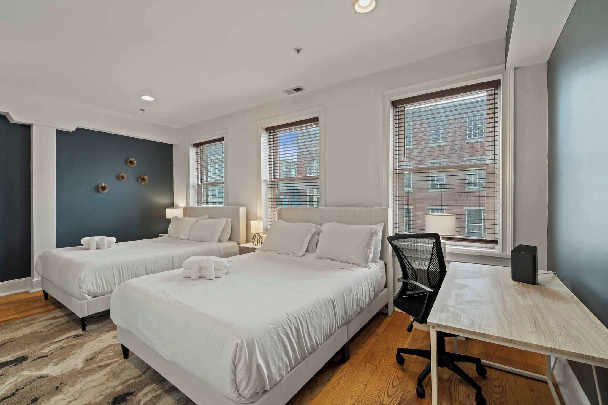 A hotel room with two beds, a desk, a chair, a lamp, and a view of a building through the window.
