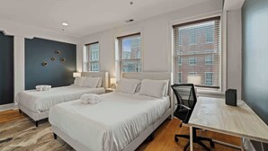 Ein Hotelzimmer mit zwei Betten, einem Schreibtisch, einem Stuhl, einer Lampe und Blick auf ein Gebäude durch das Fenster.