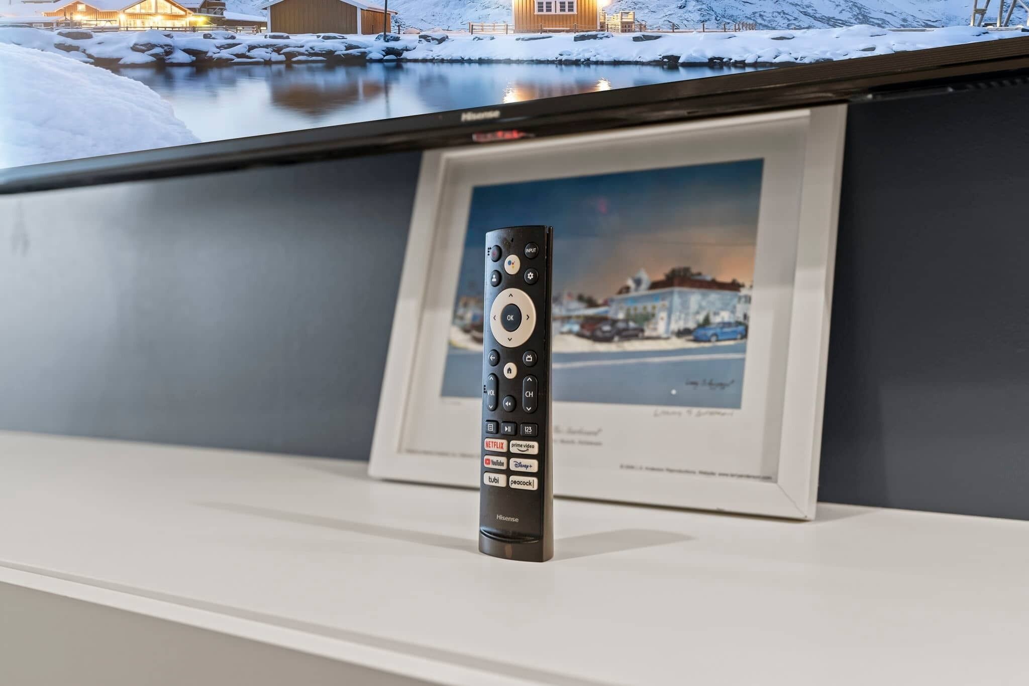 A remote control on a white surface with a framed picture of a snowy landscape in the background.