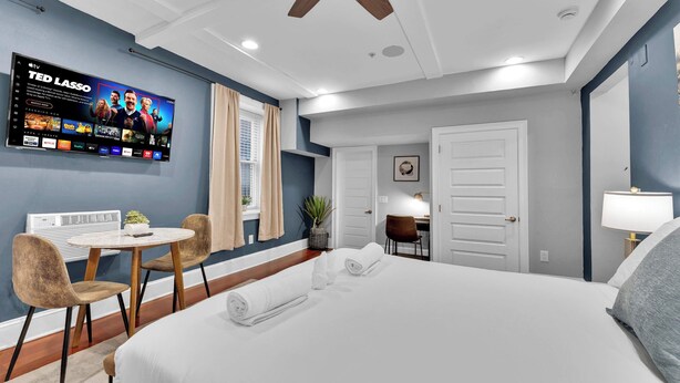 Ein Hotelzimmer mit einem großen Bett, einem Schreibtisch mit Stuhl, einem an der Wand befestigten Fernseher und einer Tür zu einem weiteren Zimmer.