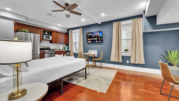 Une chambre d’hôtel moderne équipée d’une kitchenette, d’un téléviseur fixé au mur, d’un ventilateur de plafond et d’un coin repas avec une table et des chaises.