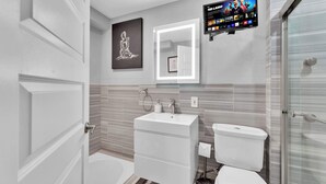 Ein modernes Badezimmer mit Waschbecken, Toilette und Dusche in Weiß. Über dem Waschbecken hängt ein gerahmtes Kunstwerk, an der Wand ist ein Flachbildfernseher angebracht.