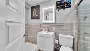 A modern bathroom with a white sink, toilet, and shower. There's a framed artwork above the sink and a flat-screen TV mounted on the wall. - Cozy Studio| Old City | 1Bed | Independence Hall (Philadelphia)
