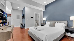 Ein modernes Hotelzimmer mit einem großen Bett, einem Schreibtisch mit Fernseher und einem durchgehenden Türspalt sichtbaren Badezimmer.