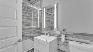 Ein modernes Badezimmer mit Duschkabine aus Glas, einem weißen Waschbecken inklusive Seifenspender, einer Toilette und einem großen Spiegel.