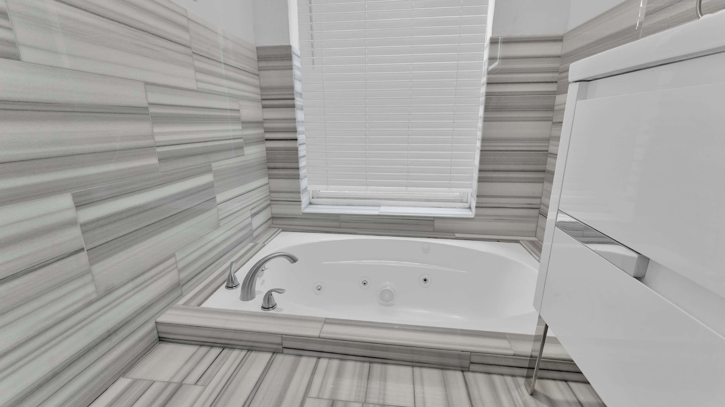 A modern bathroom with a freestanding bathtub, a window with blinds, and a tiled floor.