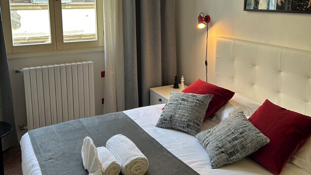 Un lit bien fait, avec des draps blancs, une couverture rayée grise et blanche, et deux serviettes enroulées. Un oreiller rouge et gris est posé sur le lit. Un radiateur blanc se trouve sous la fenêtre, et un tableau est accroché au mur.
