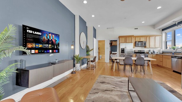 A modern kitchen with stainless steel appliances, a dining area with a long table and chairs, and a TV mounted on the wall displaying a sports channel.