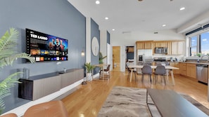 Una cocina moderna con electrodomésticos de acero inoxidable, un comedor con una mesa larga y sillas, y un televisor en la pared mostrando un canal deportivo.