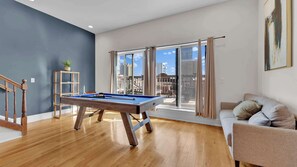 A room with a pool table, a sofa, a staircase, and a view of the city through large windows.