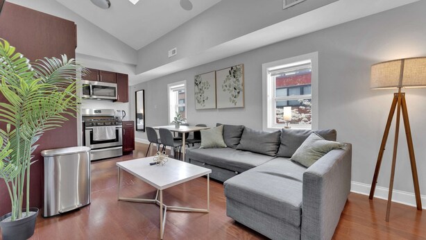 A modern living room with a grey sofa, a dining table, and a kitchen area with a microwave and oven. - Bright 1BR Retreat | Sleeps 4 | by Liberty Bell (Philadelphia)