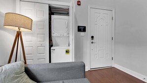 A room with a grey sofa, a lamp, a white door with a T-shaped sign, and a washing machine. - Bright 1BR Retreat | Sleeps 4 | by Liberty Bell (Philadelphia)