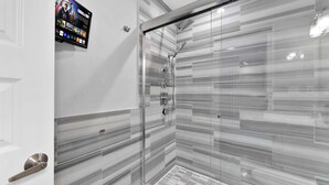 A modern bathroom with a glass shower enclosure, marble-patterned walls, and a mounted digital screen displaying content. - Bright 1BR Retreat | Sleeps 4 | by Liberty Bell (Philadelphia)
