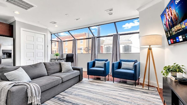 Una sala de estar moderna con un sofá gris, dos sillones azules, una lámpara de pie y un amplio ventanal con vista a la ciudad.