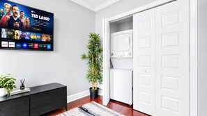 Un salon moderne avec un téléviseur à écran plat, un meuble en bois foncé, une plante en pot, un lave-linge et une porte blanche.
