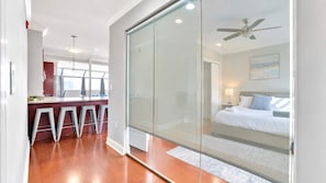 Habitación de hotel moderna con separador de vidrio, cama, ventilador de techo y vista al área de la cocina contigua.