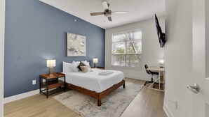 A bedroom with a bed, bedside tables, a desk, a chair, a ceiling fan, a large window with blinds, and a wall-mounted TV. - Luxury Modern 2BR Sleeps 8 | near Fair Park Dallas (Dallas)