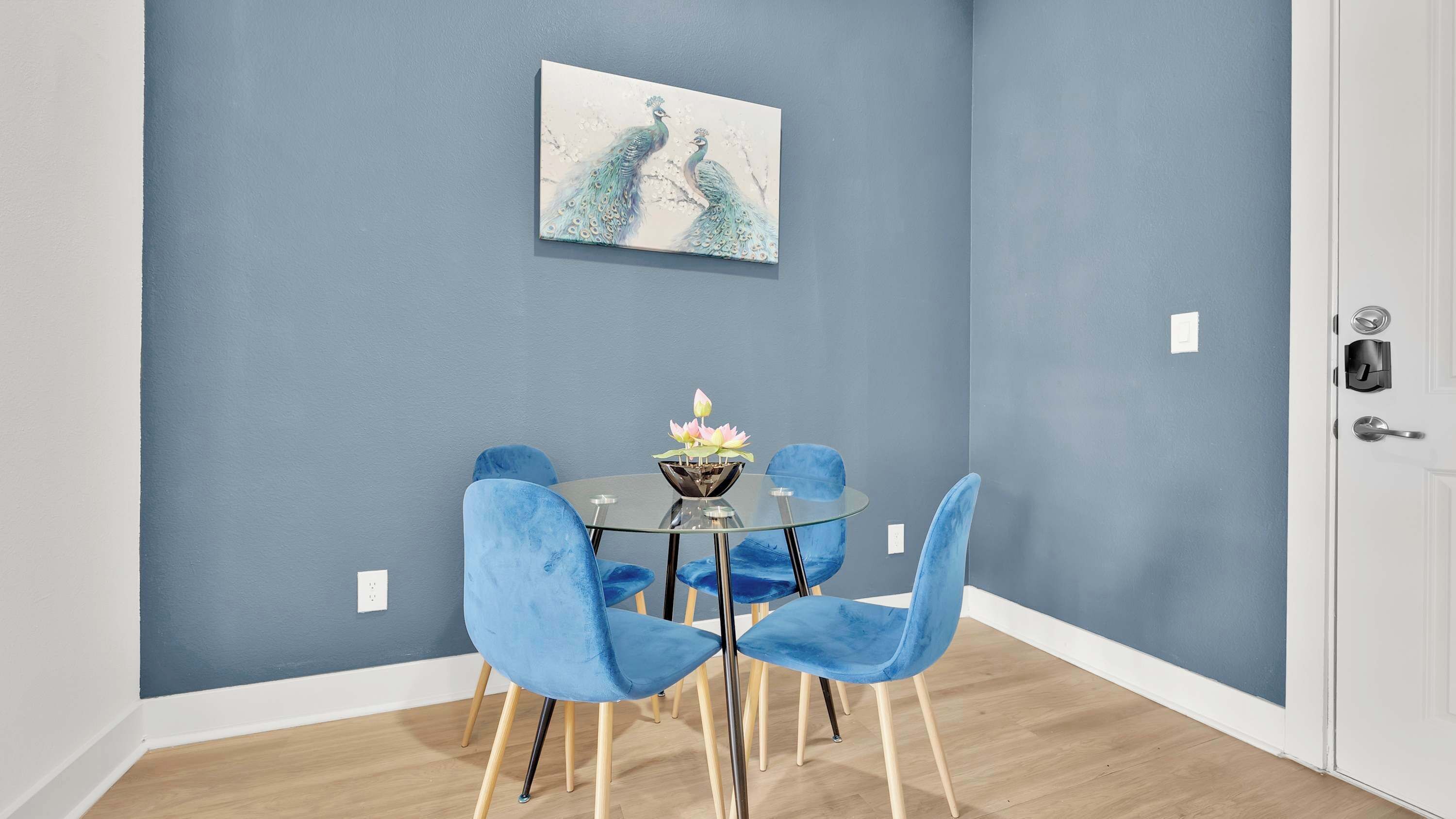 A dining area with a round glass table, four blue chairs, and a decorative bowl with lotus flowers.