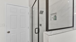 A bathroom with a white door, a glass shower enclosure, and a shelf with toiletries.