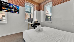 A hotel room with a large flat-screen TV mounted on the wall, a desk with a chair, a bed with white bedding, and a window with blinds. - Luxury 2BD | 4 Beds | Northern Liberties (Philadelphia)