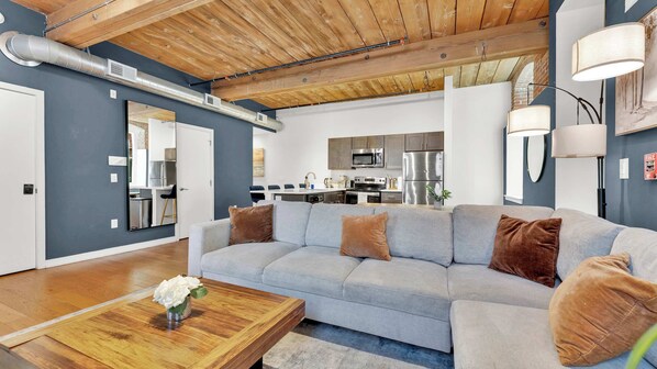 Un salon moderne avec un canapé gris, une table basse en bois et une cuisine en arrière-plan.